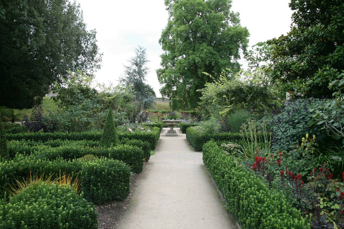 A serene garden with a central gravel path lined by neatly trimmed hedges. In the distance, a circular birdbath stands at the intersection of two paths, surrounded by lush greenery and colourful flowers. The sky is overcast, casting a soft, diffused light on the scene.